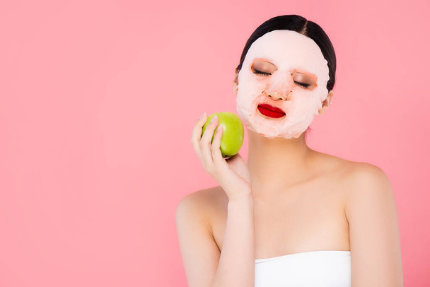 young asian woman in face mask holding green apple with closed eyes isolated on pink - Foto, imagen