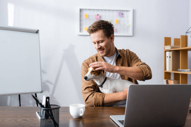 молодой бизнесмен обнимается с Джеком Расселом терьером, сидя за рабочим столом возле ноутбука и чашки кофе - Фото, изображение