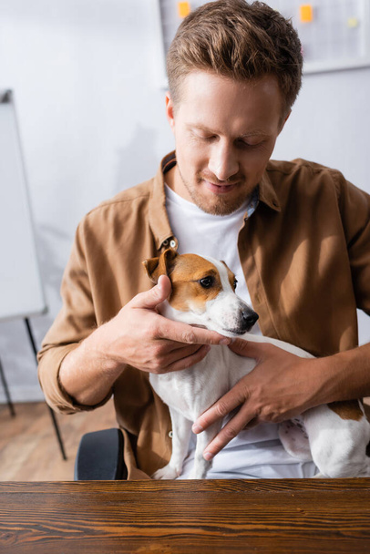 νεαρός επιχειρηματίας σε casual ρούχα κρατώντας Jack Russell Terrier σκυλί, ενώ κάθεται στο χώρο εργασίας - Φωτογραφία, εικόνα