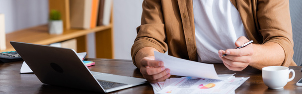 Ausgeschnittene Ansicht eines Geschäftsmannes, der mit Papieren in Laptopnähe arbeitet, horizontales Bild - Foto, Bild