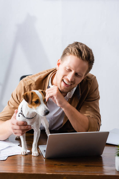 jonge zakenman in casual kleding aanraken jack russell terrier hond op het werk in de buurt laptop - Foto, afbeelding
