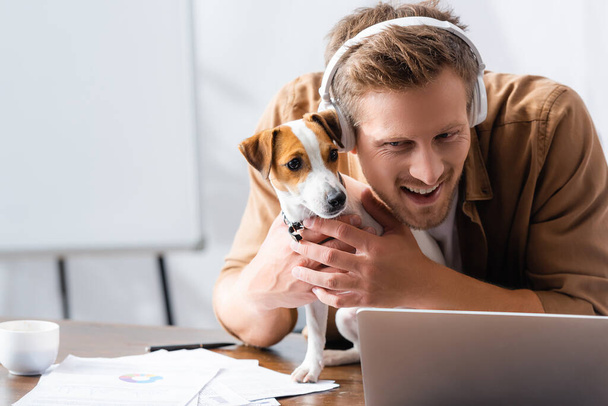 young businessman in wireless headphones cuddling jack russell terrier dog at workplace near laptop - Photo, image