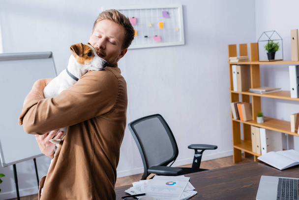 jonge zakenman houden jack russell terrier hond terwijl staan in de buurt van bureau in het kantoor - Foto, afbeelding
