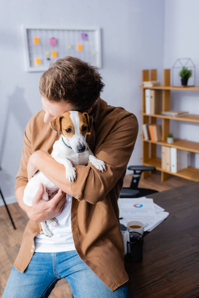 businessman in casual clothes cuddling jack russell terrier dog while sitting on office desk - Photo, Image