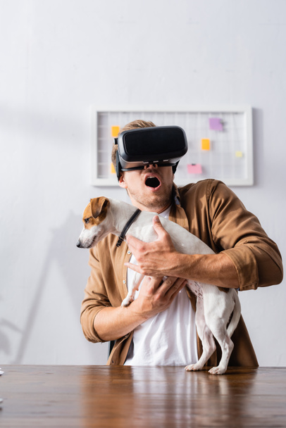 bange zakenman in VR headset houdt Jack Rassell terrier hond in het kantoor - Foto, afbeelding