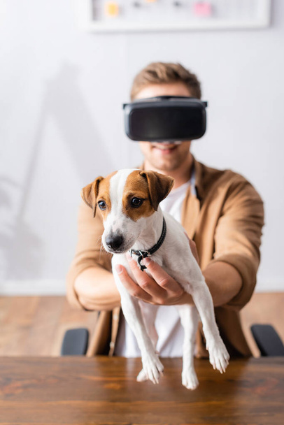 selective focus of young businessman in vr headset holding jack rassell terrier dog in office - Photo, Image