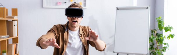 horizontal image of young businessman with open mouth using vr headset and gesturing at workplace - Photo, Image