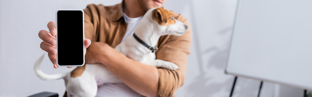 vista parcial del hombre de negocios que muestra el teléfono inteligente con la pantalla en blanco mientras sostiene el perro del terrier de Jack Russell, tiro panorámico - Foto, Imagen