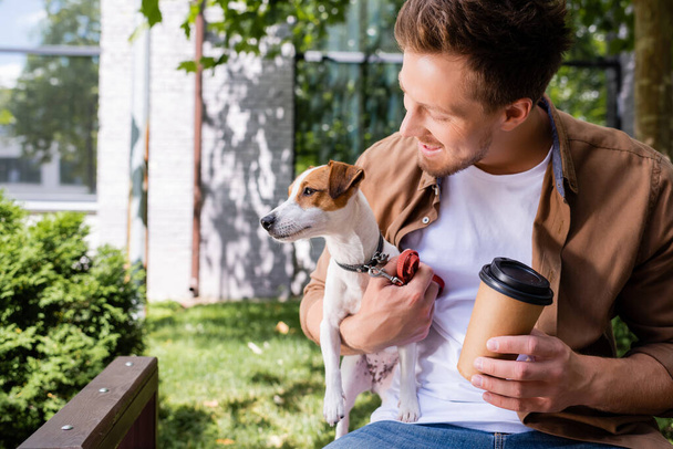 νεαρός άνδρας κρατώντας Jack Russell Terrier σκυλί, ενώ κάθεται στον πάγκο με καφέ για να πάει - Φωτογραφία, εικόνα