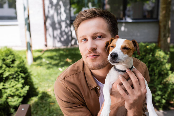 νεαρός άνδρας κοιτάζει κάμερα ενώ αγκαλιάζει Jack Russell Terrier σκυλί έξω - Φωτογραφία, εικόνα