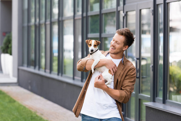 νεαρός άνδρας σε casual ρούχα κρατώντας Jack Russell τεριέ σκυλί κοντά στο κτίριο με γυάλινη πρόσοψη - Φωτογραφία, εικόνα