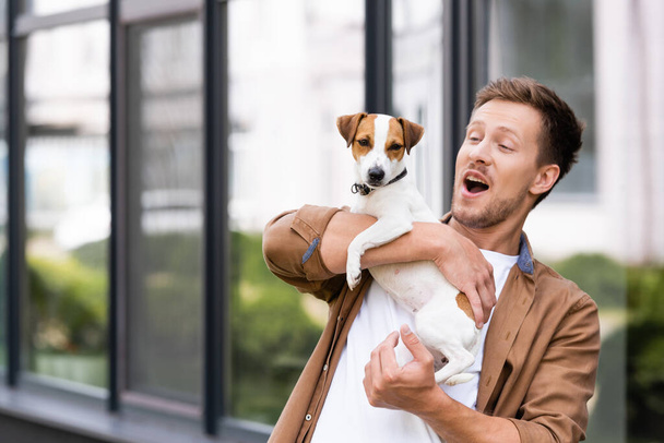 excited man with open mouth holding jack Russell terrier dog on city street - Фото, изображение