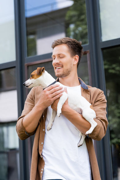 young man in casual clothes holding jack russell terrier dog on urban street - Zdjęcie, obraz