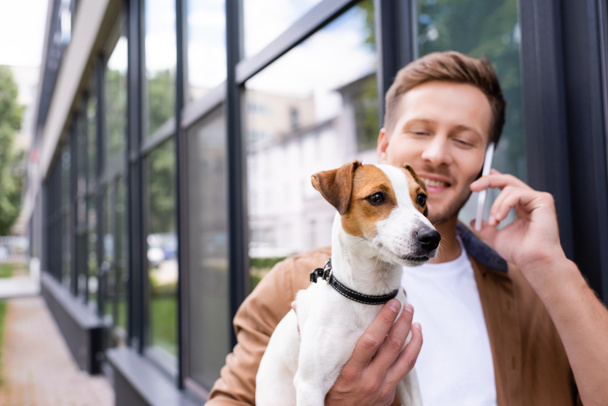 selective focus of young man holding jack russell terrier dog and talking on smartphone outdoors - Fotografie, Obrázek