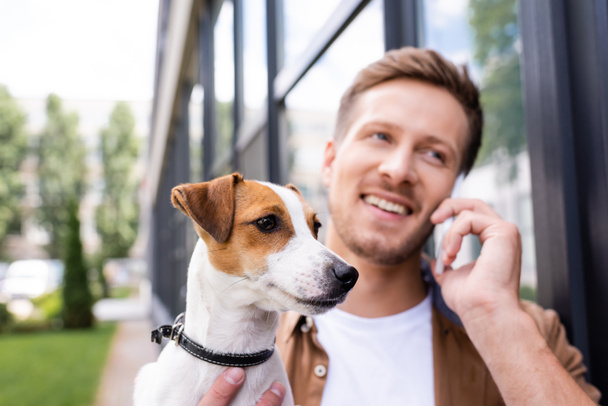 selektivní zaměření mladý muž mluví na smartphone, zatímco drží jack Russell teriér pes na ulici - Fotografie, Obrázek