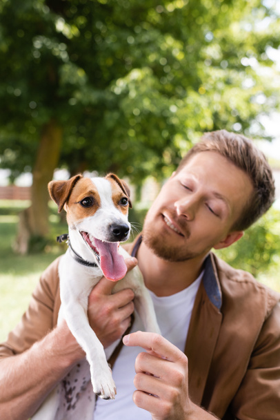 giovane uomo in possesso di jack bianco russell terrier cane con macchie marroni sulla testa nel parco - Foto, immagini