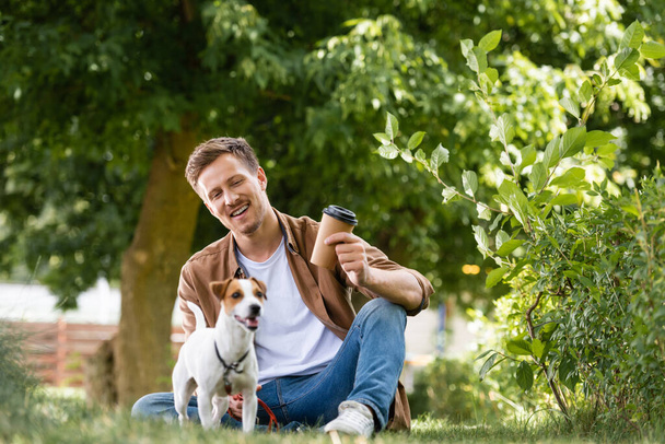 jovem homem em camisa marrom e jeans segurando copo descartável enquanto sentado na grama perto de Jack Russell terrier dog - Foto, Imagem
