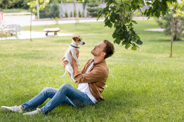 joven hombre en jeans y marrón camisa sentado en verde hierba y la celebración de jack russell terrier perro - Foto, Imagen