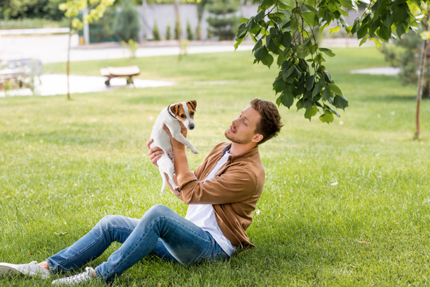 Młody mężczyzna w brązowej koszuli i dżinsach trzymający psa terrier Jacka Russella podczas siedzenia na trawniku - Zdjęcie, obraz