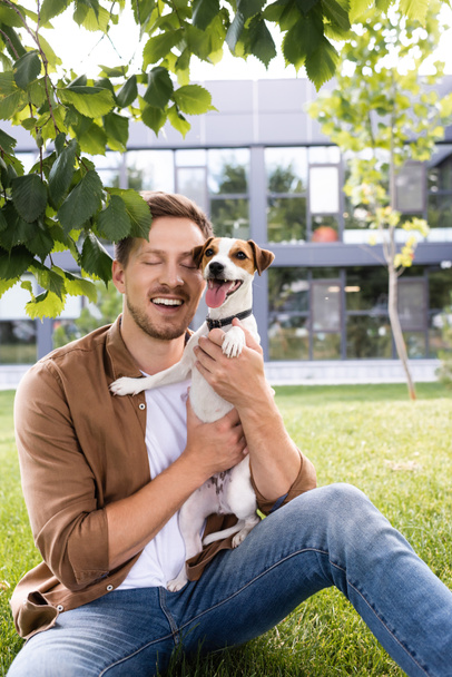 young man sitting on grass under tree branch and holding jack russell terrier dog - Photo, Image
