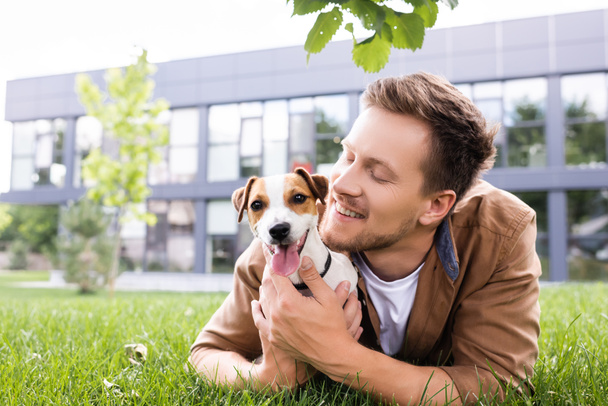 młody człowiek przytulanie Jack Russell terrier pies leżąc na zielonym trawniku  - Zdjęcie, obraz