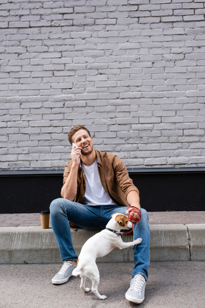 Hombre sosteniendo jack russell terrier en correa mientras habla en el teléfono inteligente cerca de café para ir en la calle urbana  - Foto, imagen