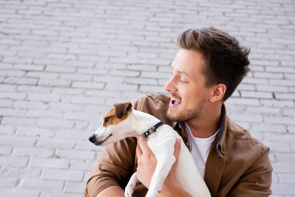 Low angle view of excited man holding jack russell terrier near facade of building  - Photo, Image