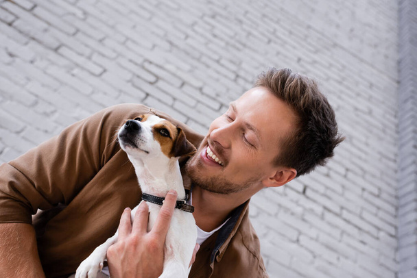Low angle view of young man embracing jack russell terrier near building - Photo, image