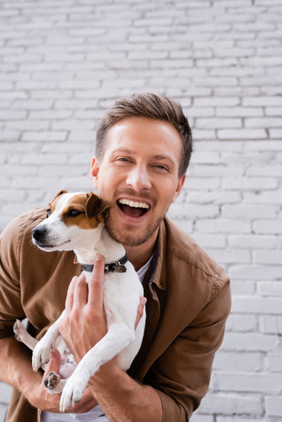Young man looking at camera while embracing jack russell terrier near facade of building  - Foto, Bild