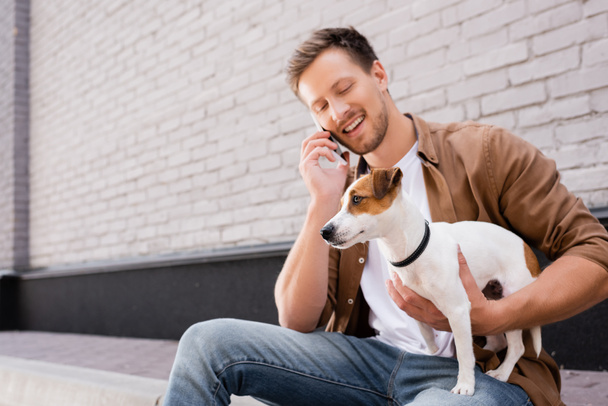 Young man talking on smartphone and hugging jack russell terrier on sidewalk near building  - 写真・画像