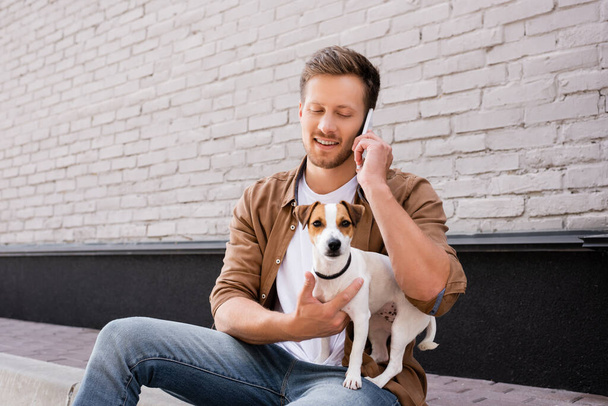 Mann umarmt Jack Russell Terrier und spricht auf Gehweg nahe Hausfassade mit Smartphone  - Foto, Bild
