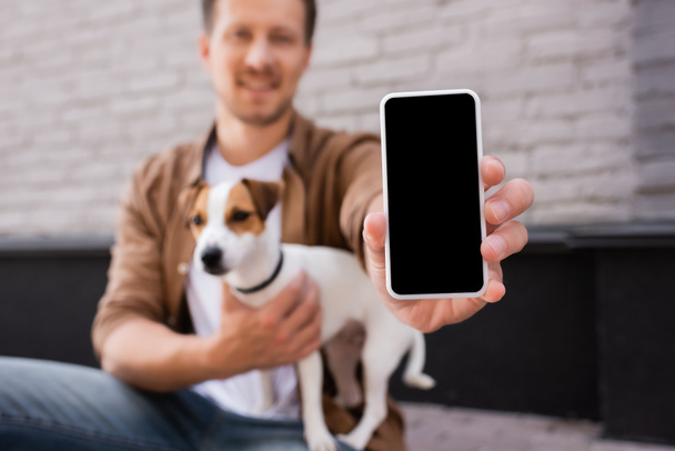 Selective focus of man showing smartphone with blank screen and embracing jack russell terrier on urban street  - Φωτογραφία, εικόνα