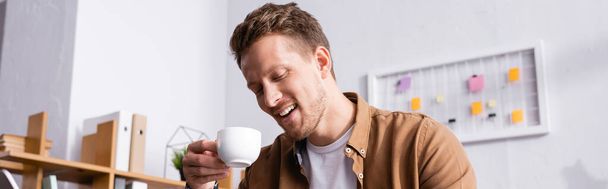 Panoramic crop of young businessman holding cup of coffee in office  - 写真・画像