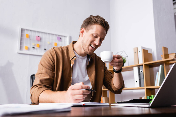Selective focus of young businessman holding cup of coffee and pen near notebook and laptop in office  - Foto, Bild