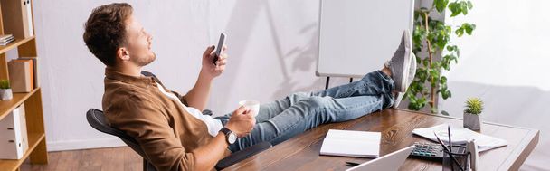Cultura horizontal de empresário usando smartphone e segurando xícara de café ao lado de artigos de papelaria na mesa de escritório  - Foto, Imagem