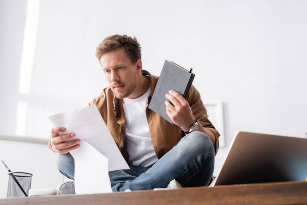 Az iratokkal és jegyzetfüzetekkel dolgozó üzletember szelektív fókusza, miközben keresztbe tett lábakkal ül az irodai asztalon  - Fotó, kép