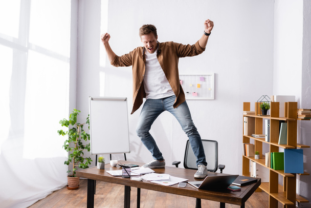Young businessman showing yes gesture while standing on table in office  - Foto, imagen