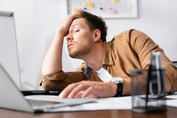 Szelektív fókusz fáradt üzletember kezét fej mellett használja laptop, miközben dolgozik az irodában  - Fotó, kép