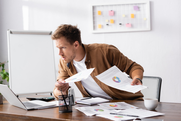 Selektivní zaměření koncentrovaného podnikatele držícího papíry a sledujícího notebook při práci v kanceláři  - Fotografie, Obrázek