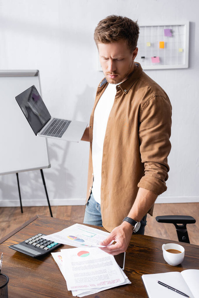 Selective focus of young businessman looking at paperwork and holding laptop near calculator and coffee in office  - Foto, imagen