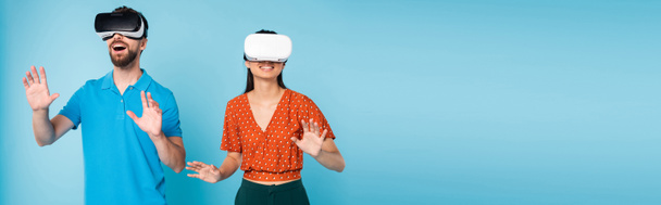 panoramic concept of excited man in polo t-shirt and woman in red blouse gesturing with hands while using vr headsets on blue - Foto, afbeelding