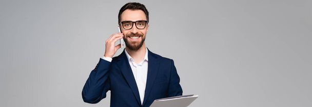 panoramic crop of bearded businessman in blue blazer talking on mobile phone and looking at camera isolated on grey - Photo, Image