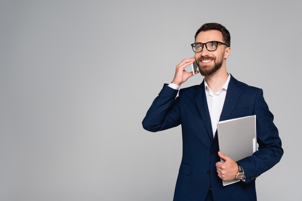 hombre de negocios barbudo en blazer azul y anteojos mirando hacia otro lado mientras habla en un teléfono inteligente aislado en gris - Foto, imagen