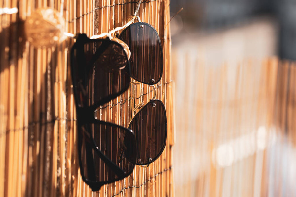 Collection of two different sunglasses models that stay on a bamboo fence in a summer day. Selective Focus - Photo, Image