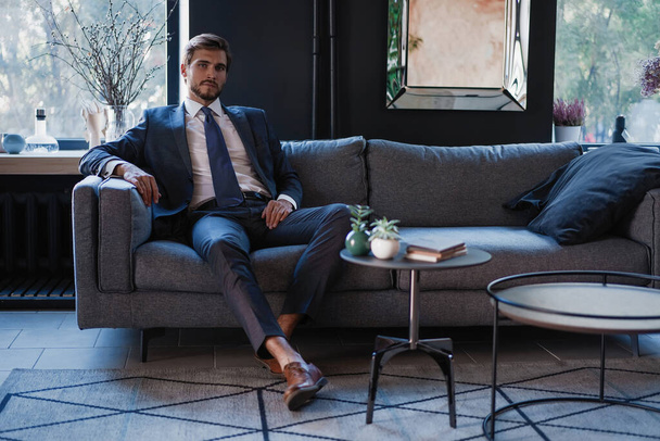 Young and charming businessman is thinking about business while sitting on the sofa in his modern office. - Foto, immagini