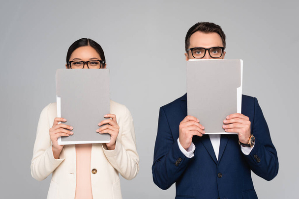 interracial couple of businesspeople in eyeglasses obscuring faces with folders isolated on grey - 写真・画像