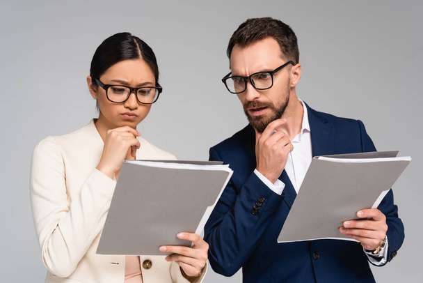 thoughtful interracial business partners in eyeglasses touching chins while looking at documents isolated on grey - Photo, Image