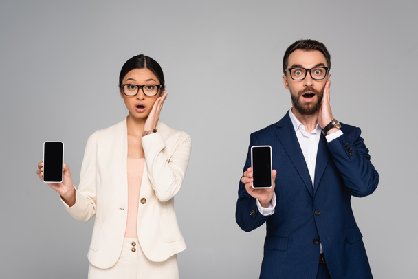 sorprendido pareja interracial de colegas de negocios tocando caras mientras sostiene los teléfonos inteligentes con pantalla en blanco aislado en gris - Foto, imagen