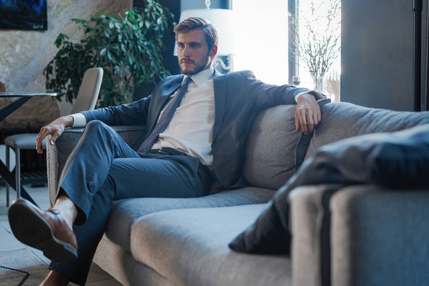 Young and charming businessman is thinking about business while sitting on the sofa in his modern office. - Φωτογραφία, εικόνα
