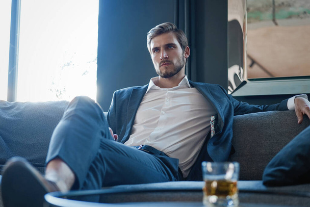 Mladý a okouzlující podnikatel přemýšlí o podnikání, zatímco sedí na pohovce ve své moderní kanceláři. - Fotografie, Obrázek
